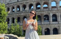 A smiling young woman making a W sign in front of Roman ruins.