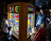 Children and adults looking at a large lite brite type structure with glowing pegs spelling out science.