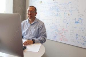 John Yin in his office looking at his computer. A well marked whiteboard is behind him.