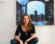 A woman sits crosslegged in front a painting with a person standing in a doorway.