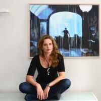 A woman sits crosslegged in front a painting with a person standing in a doorway.