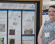A woman stands in front a scientific poster with short brown hair and sleeveless, patterned top.