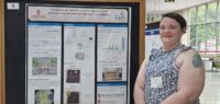 A woman stands in front a scientific poster with short brown hair and sleeveless, patterned top.
