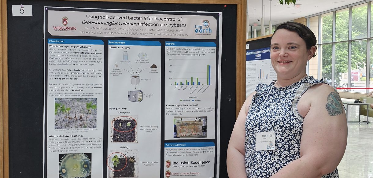 A woman stands in front a scientific poster with short brown hair and sleeveless, patterned top.