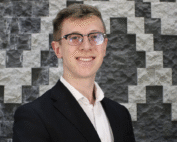 Ben Lukszys standing in front of a grey and white patterned brick wall wearing a dark suit and glasses.