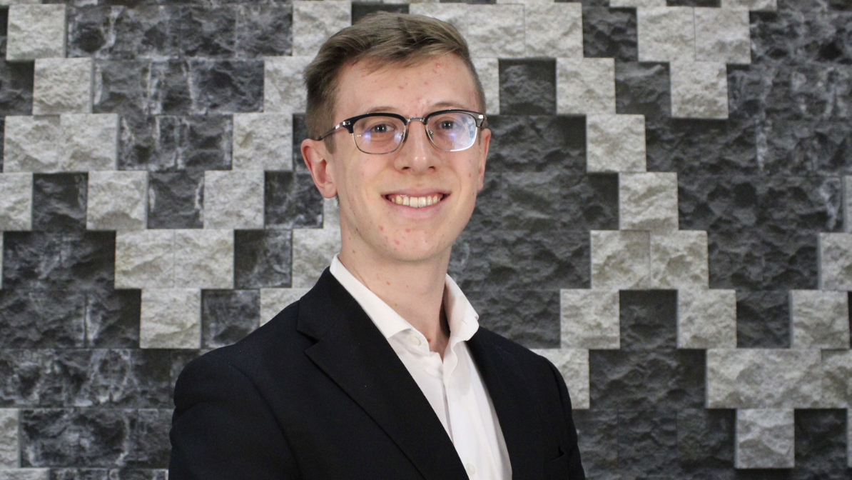 Ben Lukszys standing in front of a grey and white patterned brick wall wearing a dark suit and glasses.