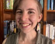 A woman is smiling in front of a stocked bookshelf wearing interesting earrings.