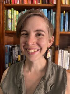 A woman is smiling in front of a stocked bookshelf wearing interesting earrings.