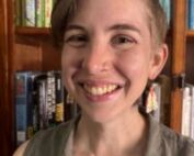 A woman is smiling in front of a stocked bookshelf wearing interesting earrings.