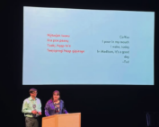Two well dressed indigenous people stand by a podium on a dark stage with a ho-chunk language poem on a screen