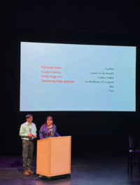 Two well dressed indigenous people stand by a podium on a dark stage with a ho-chunk language poem on a screen