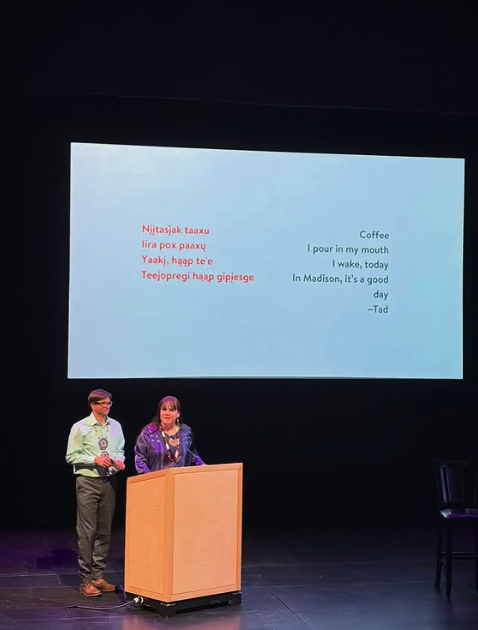 Two well dressed indigenous people stand by a podium on a dark stage with a ho-chunk language poem on a screen